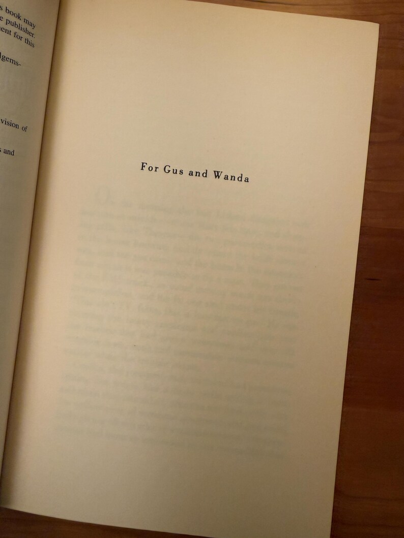Op de afbeelding: Open boek met de tekst "For Gus and Wanda" in zwarte letters op een cr&egrave;mekleurige pagina. Het boek ligt op een houten oppervlak.