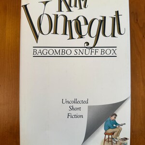 May include: A hardcover book titled "Bagombo Snuff Box" by Kurt Vonnegut. The cover is white with black and gold lettering. The book features an illustration of a person sitting on a stool reading.