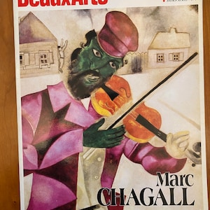 May include: A magazine cover featuring a painting by Marc Chagall. The painting depicts a man with a green face playing a violin. The man is wearing a purple jacket and a red hat. The background is a simple landscape with two houses.