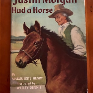 Puede incluir: Portada del libro "Justin Morgan Had a Horse" de Marguerite Henry, ilustrado por Wesley Dennis. Muestra un caballo marrón y un hombre con sombrero y chaleco. El título está en letras rojo oscuro.
