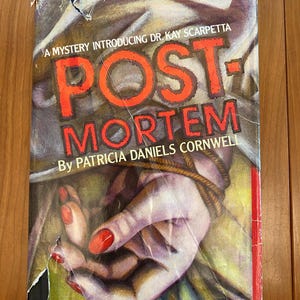 May include: A paperback book titled "Postmortem" by Patricia Daniels Cornwell. The cover features a hand tied with rope, with red nail polish. The title is in large, red, block letters.