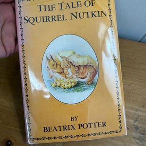Może przedstawiać: Vintage egzemplarz książki "The Tale of Squirrel Nutkin" autorstwa Beatrix Potter. Książka ma jasno pomarańczową okładkę z tytułem w czarnym tekście i okrągłą ilustracją wiewiórek. Okładka jest obramowana ozdobną bordiurą.
