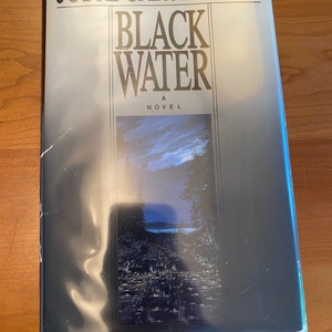 Könnte beinhalten: Ein Hardcover-Buch mit dem Titel "Black Water" von Joyce Carol Oates. Der Buchumschlag zeigt ein dunkelblaues und schwarzes Bild eines Sees und Bäume bei Nacht.