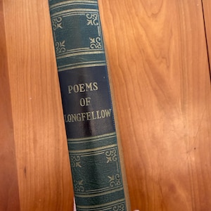 Puede incluir: Un libro antiguo titulado "Poems of Longfellow". El libro tiene una cubierta verde oscuro con detalles decorativos dorados y un lomo azul marino. El título está en letras doradas. El libro se sostiene contra una superficie de madera.
