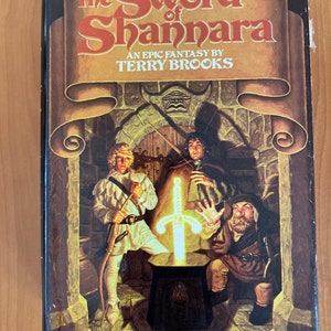 Puede incluir: Una portada de libro con una ilustración de fantasía de tres personas de pie en una habitación de piedra. El título del libro es "The Sword of Shannara" y el autor es "Terry Brooks".