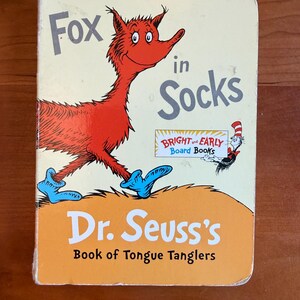Può includere: Un libro per bambini intitolato "Fox in Socks" di Dr. Seuss. La copertina presenta un personaggio volpe rossa con calzini blu, con il titolo in grandi lettere giocose. Il libro fa parte della serie "Bright and Early Board Books".