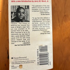 Op de afbeelding: Achterkant van een paperback boek met een portret en tekst. De witte omslag heeft zwarte tekst, inclusief recensies en het logo van de uitgever. Titel en auteur van het boek zijn niet zichtbaar. De omslag is licht beschadigd.