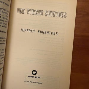 Op de afbeelding: De titelpagina van "The Virgin Suicides" van Jeffrey Eugenides. De titel staat in een zwarte, vette letter. De naam van de auteur staat eronder. Het Warner Books-logo en "A Time Warner Company" staan onderaan. De pagina's zijn cr&egrave;mekleurig.