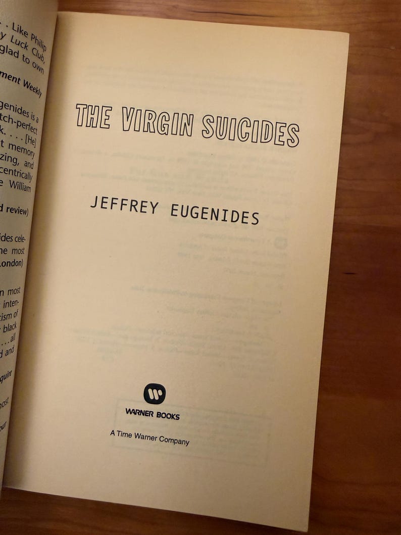 Op de afbeelding: De titelpagina van "The Virgin Suicides" van Jeffrey Eugenides. De titel staat in een zwarte, vette letter. De naam van de auteur staat eronder. Het Warner Books-logo en "A Time Warner Company" staan onderaan. De pagina's zijn cr&egrave;mekleurig.