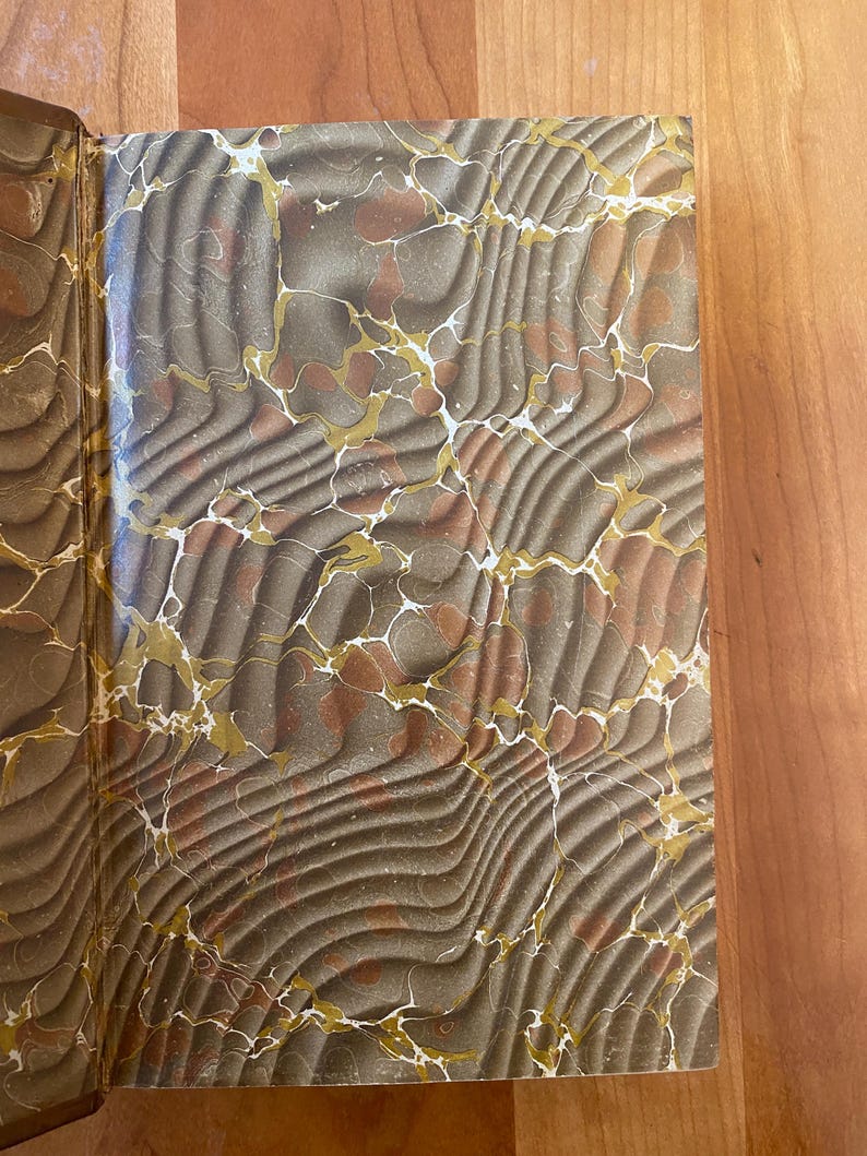 Pu&ograve; includere: Primo piano delle controguardie marmorizzate di un libro in tonalit&agrave; di marrone, oro e bianco. Il motivo marmoreo &egrave; intricato e vorticoso.
