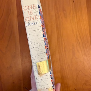 May include: A vintage book titled "One Is One" by Picard. The book's spine is white with red, blue, and gold accents. A gold-coloured sticker is affixed to the spine. The book is held against a wooden surface.