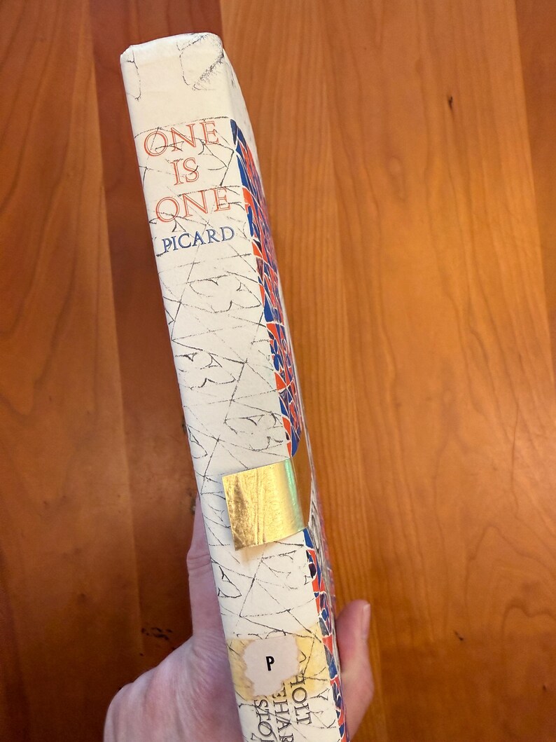 May include: A vintage book titled "One Is One" by Picard. The book's spine is white with red, blue, and gold accents. A gold-coloured sticker is affixed to the spine. The book is held against a wooden surface.