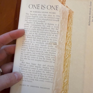 May include: A vintage book titled "One Is One" by Barbara Leonie Picard. The book's cover is partially visible, with text describing the story's setting in 14th-century England. The jacket is designed by Christine Freisinger.