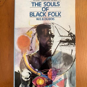 Könnte beinhalten: Ein Taschenbuch mit dem Titel "THE SOULS OF BLACK FOLK" von W.E.B. DuBois. Das Cover zeigt ein Porträt eines schwarzen Mannes mit abstrakten Kunstelementen, darunter ein Regenbogen, ein Auge und eine Flagge.