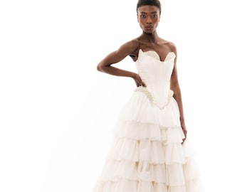 Puede incluir: Un vestido de novia marfil sin tirantes con una falda con volantes en capas y una larga cola. El corpiño está adornado con delicadas cuentas. El vestido está diseñado con una silueta ajustada y un escote corazón.