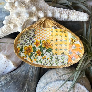 May include: A gold-painted seashell dish with a floral pattern in shades of yellow, orange, and white. The dish is decorated with a honeycomb pattern and a small bee.