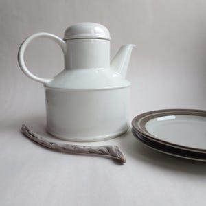 Könnte beinhalten: Eine weiße Keramik-Teekanne mit gebogenem Griff und Deckel, neben einem Stapel Teller mit braunen und weißen Rändern. Ein Stück Treibholz befindet sich im Vordergrund. Die Objekte befinden sich auf einer weißen Oberfläche.