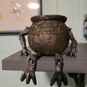 May include: A black and gold cauldron with a detailed design sits on a shelf. The cauldron has four legs that resemble clawed feet.