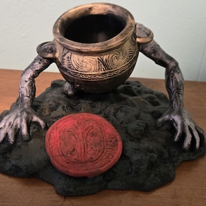 May include: A decorative, fantasy-themed sculpture featuring a cauldron-shaped vessel with arms and hands. The cauldron is bronze-colored with etched designs. It rests on a dark, textured base with a red, circular emblem. The sculpture is on a wooden surface.