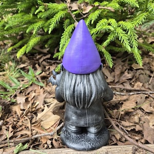 May include: A garden gnome statue with a purple pointed hat and a gray coat. The gnome is facing away from the camera, standing on a wooden surface with a backdrop of green foliage and fallen leaves.