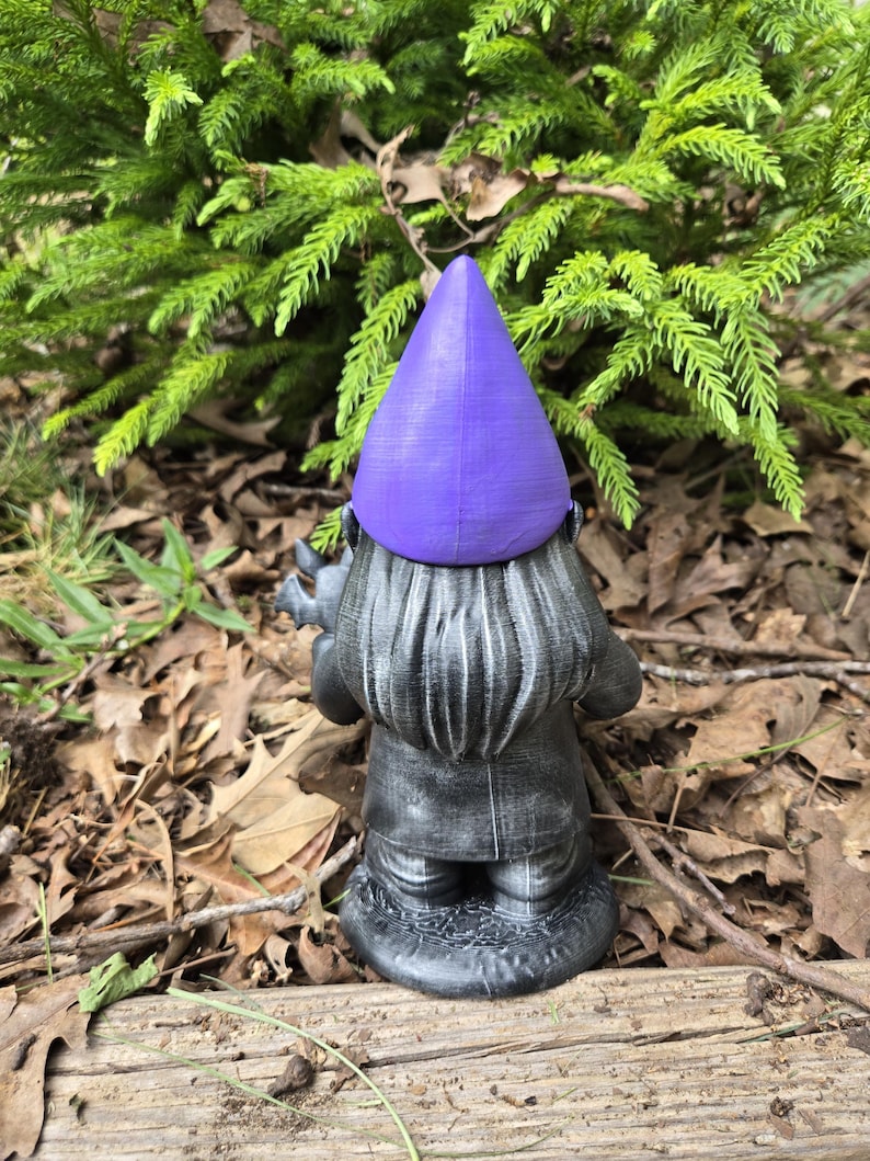 May include: A garden gnome statue with a purple pointed hat and a gray coat. The gnome is facing away from the camera, standing on a wooden surface with a backdrop of green foliage and fallen leaves.