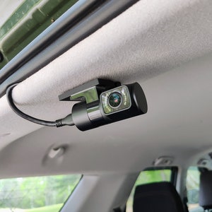 May include: A black dash cam mounted to the ceiling of a car. The camera has a lens that is pointed forward and a small screen on the side. The camera is connected to a power source by a black cable.