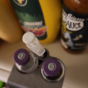 Puede incluir: Primer plano de un soporte met&aacute;lico con dos viales con tapas moradas y una jeringa de pl&aacute;stico transparente. Una botella de mostaza amarilla y una botella de salsa en el fondo.
