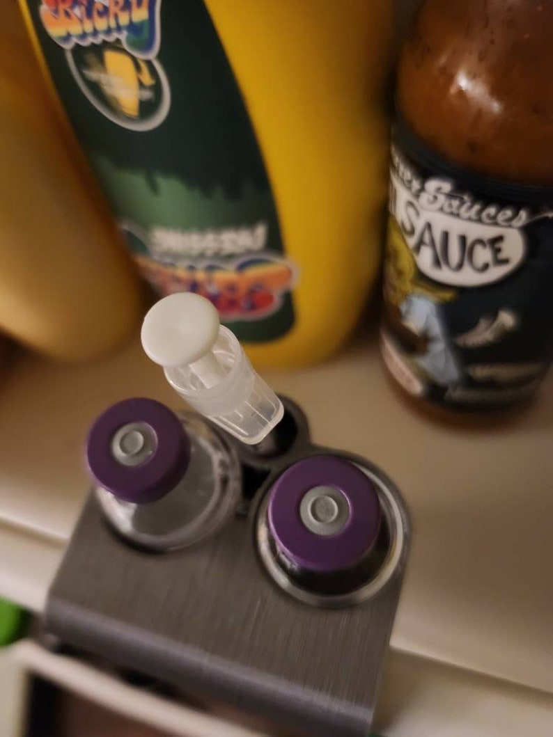 Puede incluir: Primer plano de un soporte met&aacute;lico con dos viales con tapas moradas y una jeringa de pl&aacute;stico transparente. Una botella de mostaza amarilla y una botella de salsa en el fondo.
