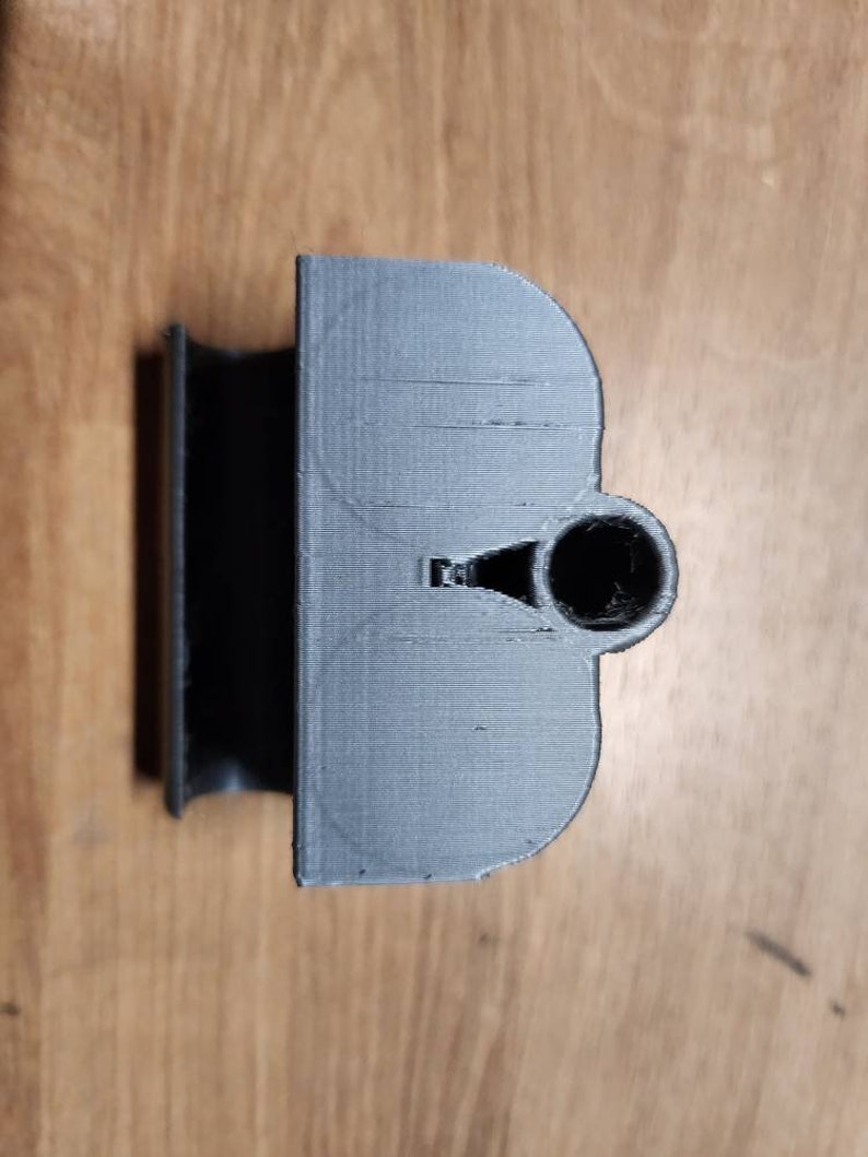 Puede incluir: Un objeto gris impreso en 3D con una parte superior redondeada y un agujero cil&iacute;ndrico. El objeto tiene una base rectangular y un dise&ntilde;o curvo semicircular. La superficie tiene l&iacute;neas de capa visibles.