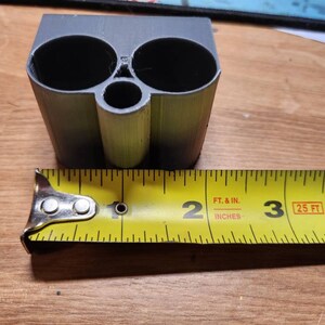 Puede incluir: Un objeto gris impreso en 3D con dos grandes aberturas circulares y una abertura m&aacute;s peque&ntilde;a. El objeto mide aproximadamente 5 cm de ancho, medido con una cinta m&eacute;trica amarilla.