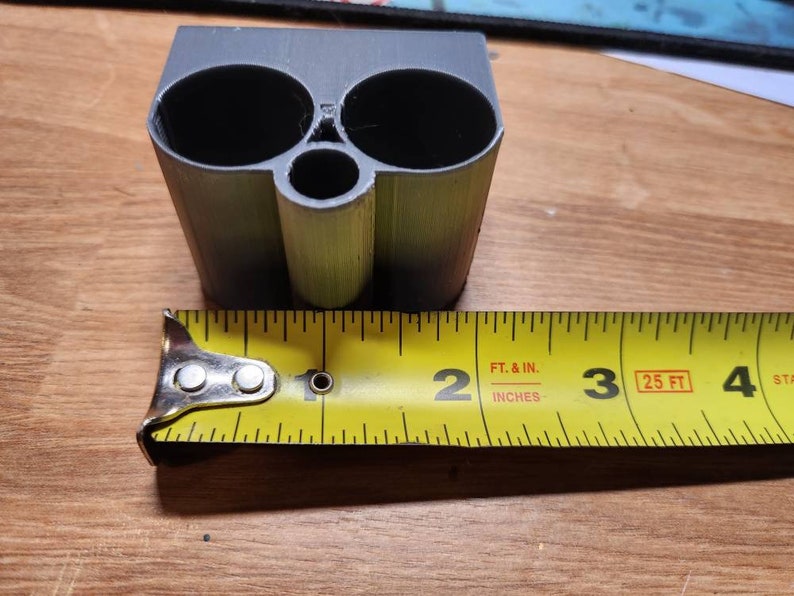 Puede incluir: Un objeto gris impreso en 3D con dos grandes aberturas circulares y una abertura m&aacute;s peque&ntilde;a. El objeto mide aproximadamente 5 cm de ancho, medido con una cinta m&eacute;trica amarilla.