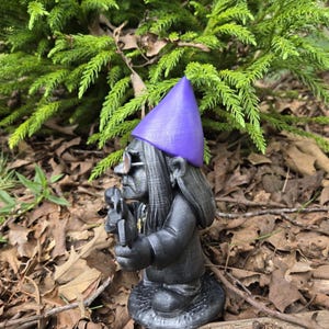 May include: A black garden gnome statue with a purple hat and glasses, holding a small tool. The gnome has long hair and is standing on a textured base. The background features green foliage and brown leaves.