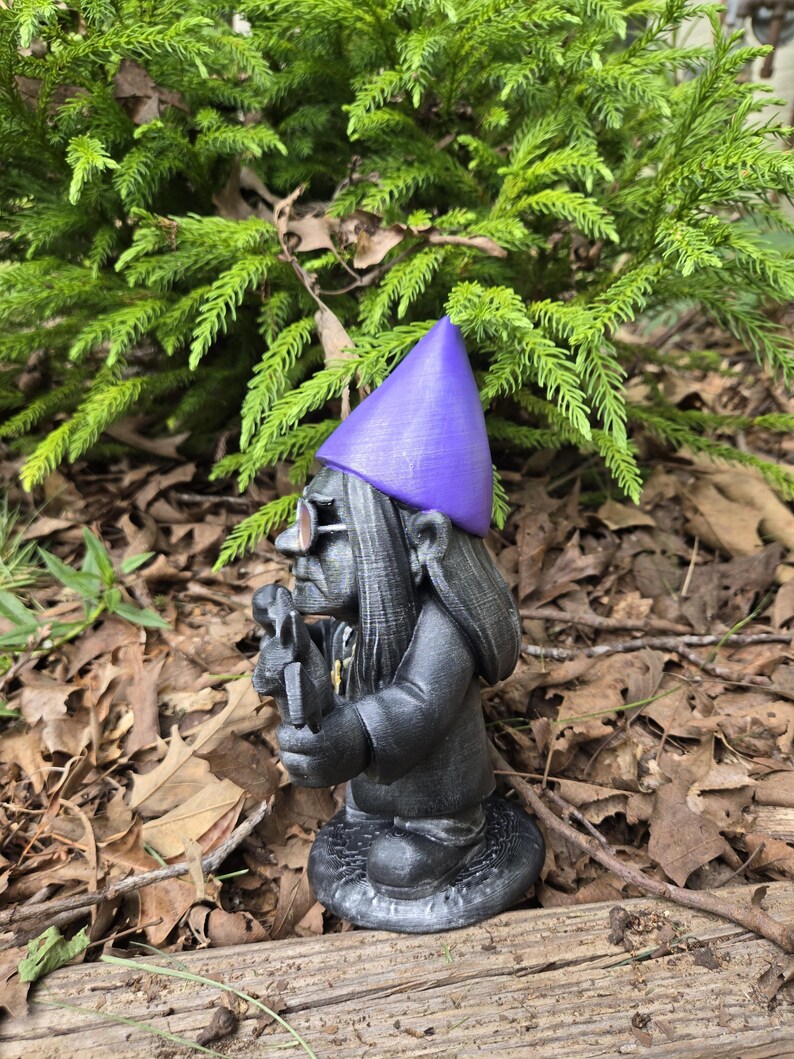 May include: A black garden gnome statue with a purple hat and glasses, holding a small tool. The gnome has long hair and is standing on a textured base. The background features green foliage and brown leaves.