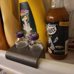 Puede incluir: Un organizador gris impreso en 3D sostiene dos peque&ntilde;os viales de vidrio con tapas moradas. Una botella de salsa picante con una etiqueta de dibujos animados y una botella amarilla tambi&eacute;n son visibles en el refrigerador.