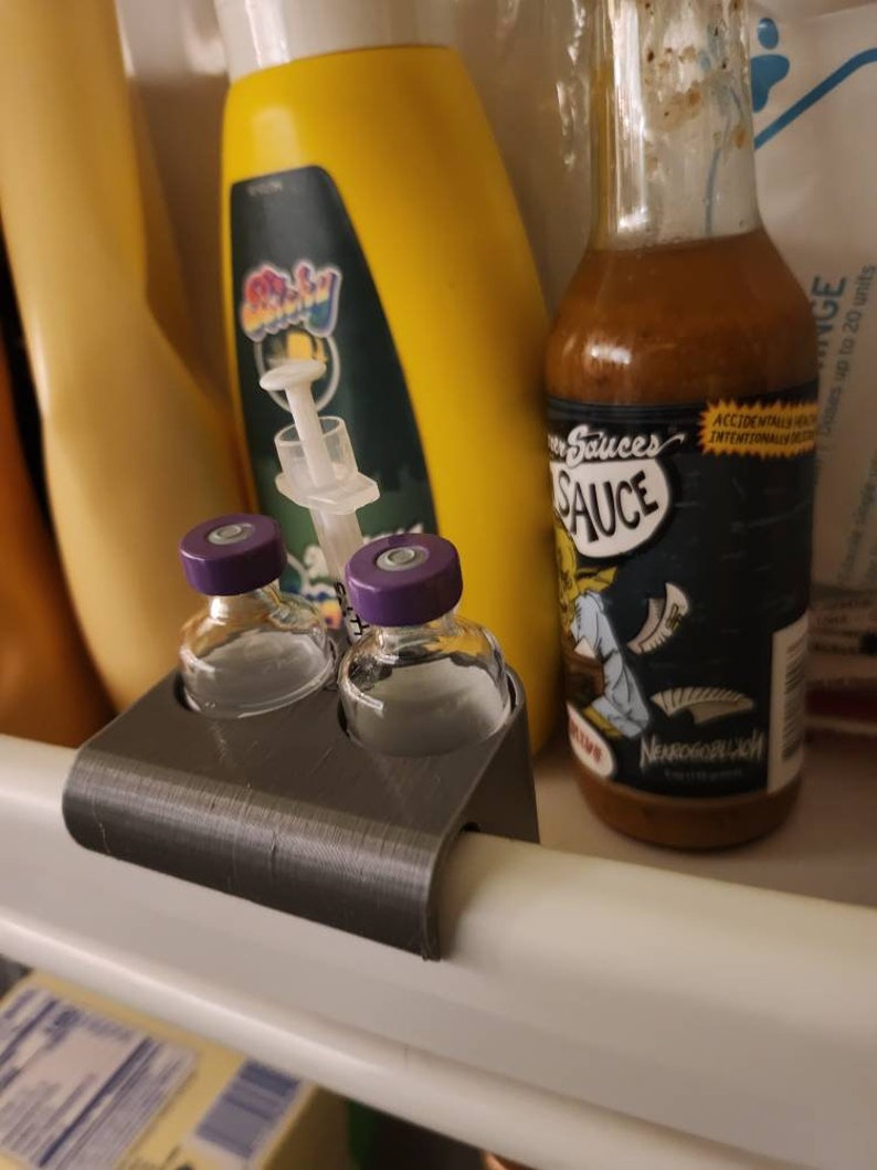 Puede incluir: Un organizador gris impreso en 3D sostiene dos peque&ntilde;os viales de vidrio con tapas moradas. Una botella de salsa picante con una etiqueta de dibujos animados y una botella amarilla tambi&eacute;n son visibles en el refrigerador.
