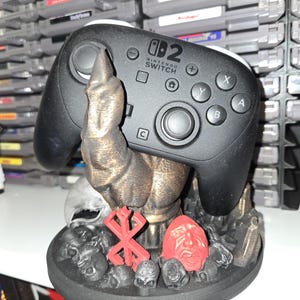May include: Black Nintendo Switch Pro controller resting on a detailed, bronze-colored stand. The stand features a red symbol, a red face, and several small skulls. The controller has the Nintendo Switch logo.