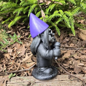 May include: A gray garden gnome statue with a purple pointed hat and glasses, holding a microphone. The gnome is standing on a round base with a textured design. The background features green foliage and fallen leaves.