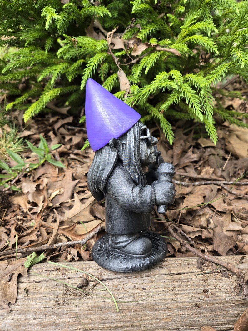 May include: A gray garden gnome statue with a purple pointed hat and glasses, holding a microphone. The gnome is standing on a round base with a textured design. The background features green foliage and fallen leaves.