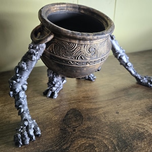 May include: A gold cauldron with a decorative design sits on three skeletal legs. The cauldron is made of ceramic and has a dark brown glaze. The legs are made of a light gray material and have a rough texture.