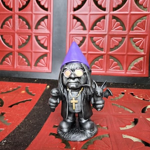May include: A dark gray gnome figurine with a purple hat, round glasses, and a black coat. The gnome holds a microphone in one hand and a bat in the other. A gold cross hangs from a chain around its neck. The figurine is on a red surface.