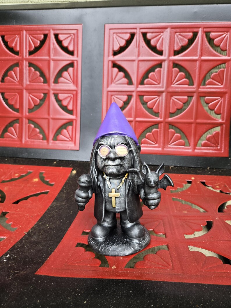 May include: A dark gray gnome figurine with a purple hat, round glasses, and a black coat. The gnome holds a microphone in one hand and a bat in the other. A gold cross hangs from a chain around its neck. The figurine is on a red surface.
