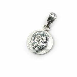 May include: Silver pendant featuring a round coin with a detailed profile of a person wearing a helmet. The pendant has a simple bail.