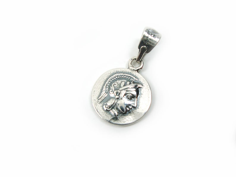 May include: Silver pendant featuring a round coin with a detailed profile of a person wearing a helmet. The pendant has a simple bail.