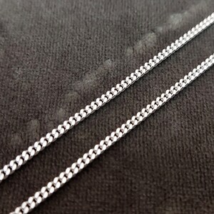 K&ouml;nnte beinhalten: Eine silberne Kettenkette im Curb-Link-Stil. Die Kette liegt flach auf einer dunkelgrauen Oberfl&auml;che.