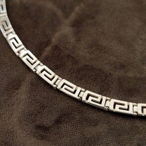 May include: A silver chain necklace with a Greek key pattern.