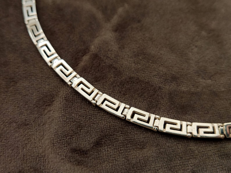 May include: A silver chain necklace with a Greek key pattern.