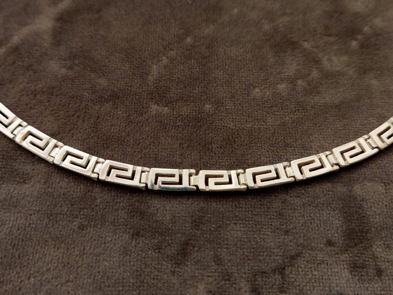 May include: A silver chain necklace with a Greek key pattern.