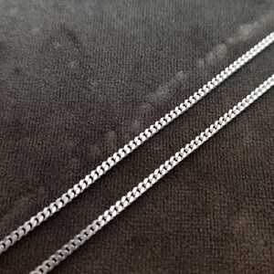 K&ouml;nnte beinhalten: Eine silberne Kettenkette im Curb-Link-Stil. Die Kette liegt flach auf einer dunkelgrauen Stoffoberfl&auml;che.