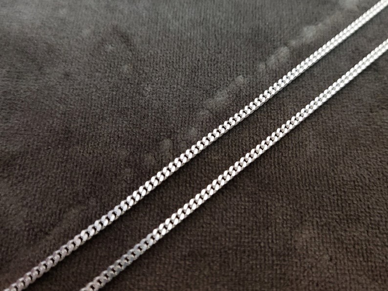K&ouml;nnte beinhalten: Eine silberne Kettenkette im Curb-Link-Stil. Die Kette liegt flach auf einer dunkelgrauen Stoffoberfl&auml;che.