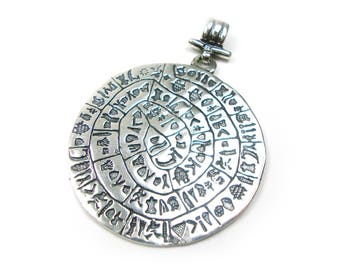 May include: Silver pendant with a circular design featuring a spiral of ancient symbols and characters. The pendant has a loop at the top for attaching to a chain.